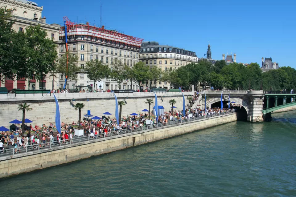 A large crowd gathers on the banks of the River Seine on a sunny day in Paris.